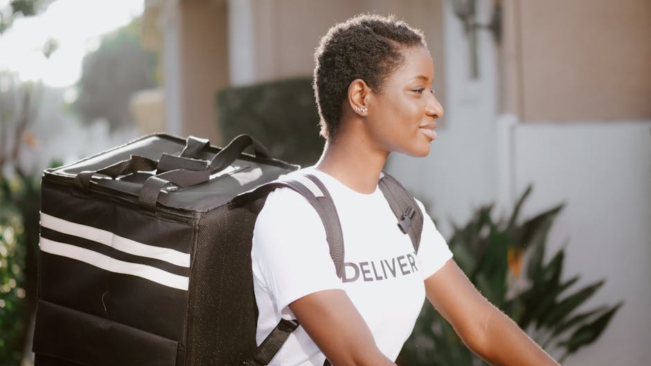 A young woman with short, curly hair and a calm expression is standing outdoors in natural sunlight, wearing a white t-shirt with the word 'DELIVER' printed on it and carrying a black insulated delive