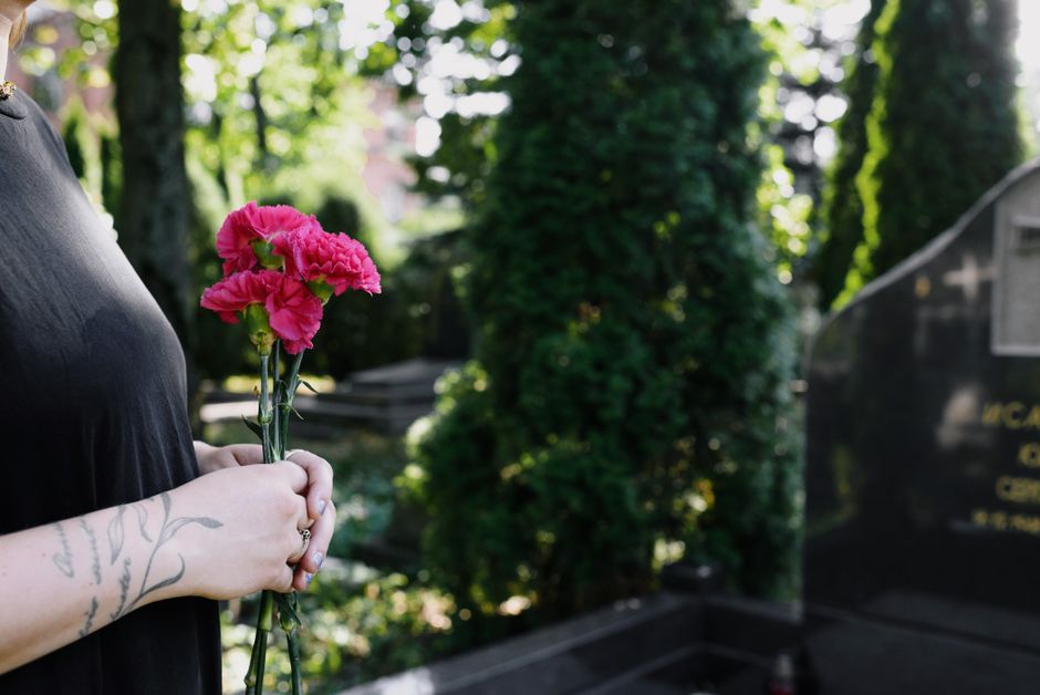 Close-up of a person holding a small bouquet of pink carnations with ruffled petals, wrapped in clear cellophane, against a background of greenery and a cemetery setting. The person is wearing a black