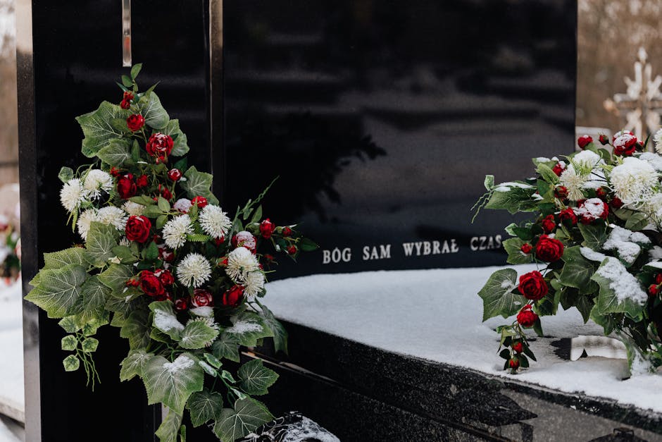 A floral arrangement placed on a black granite tombstone, featuring a mixture of red roses, white chrysanthemums, and white anemones, complemented by lush green ivy and holly leaves, some of which hav